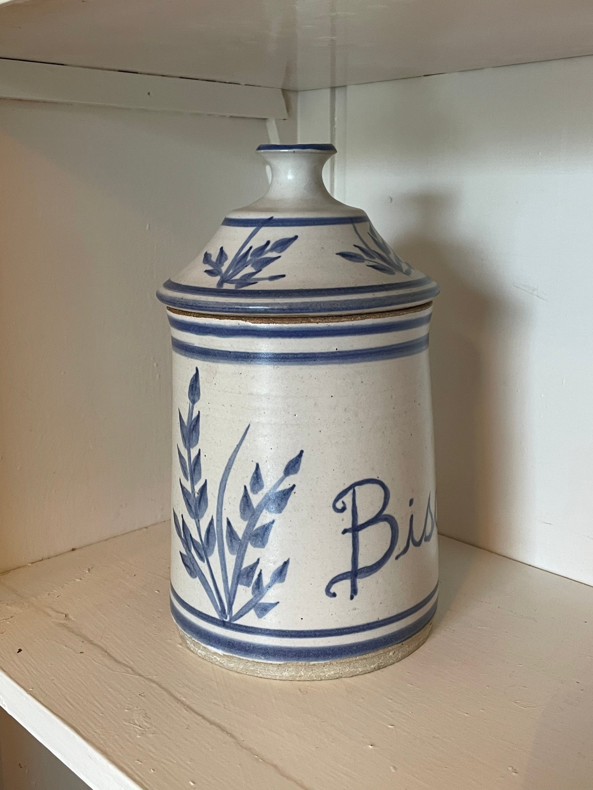 A vintage ceramic lidded storage jar with blue floral and script lettering on a cream-colored stoneware body, displayed on a wooden shelf.