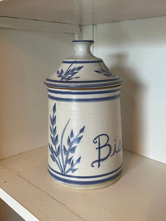 A vintage ceramic lidded storage jar with blue floral and script lettering on a cream-colored stoneware body, displayed on a wooden shelf.