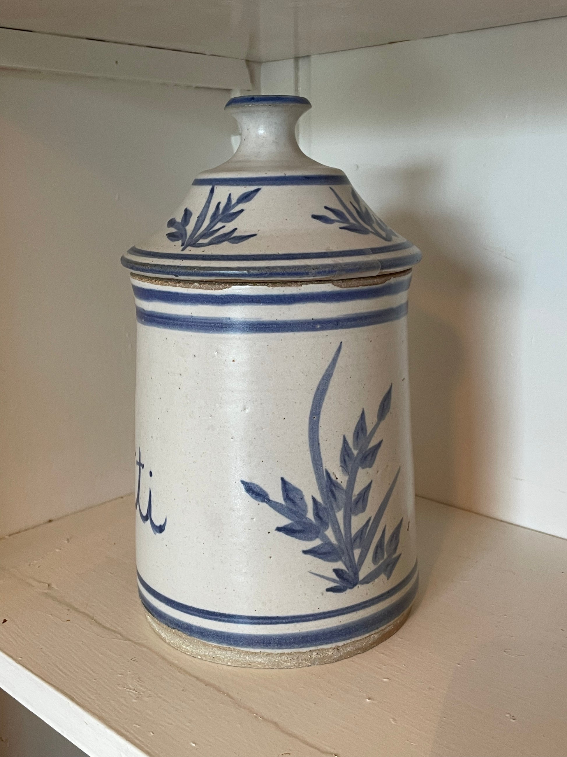 A vintage ceramic lidded canister with a cream-colored glaze and blue floral motifs, displayed on a wooden shelf.