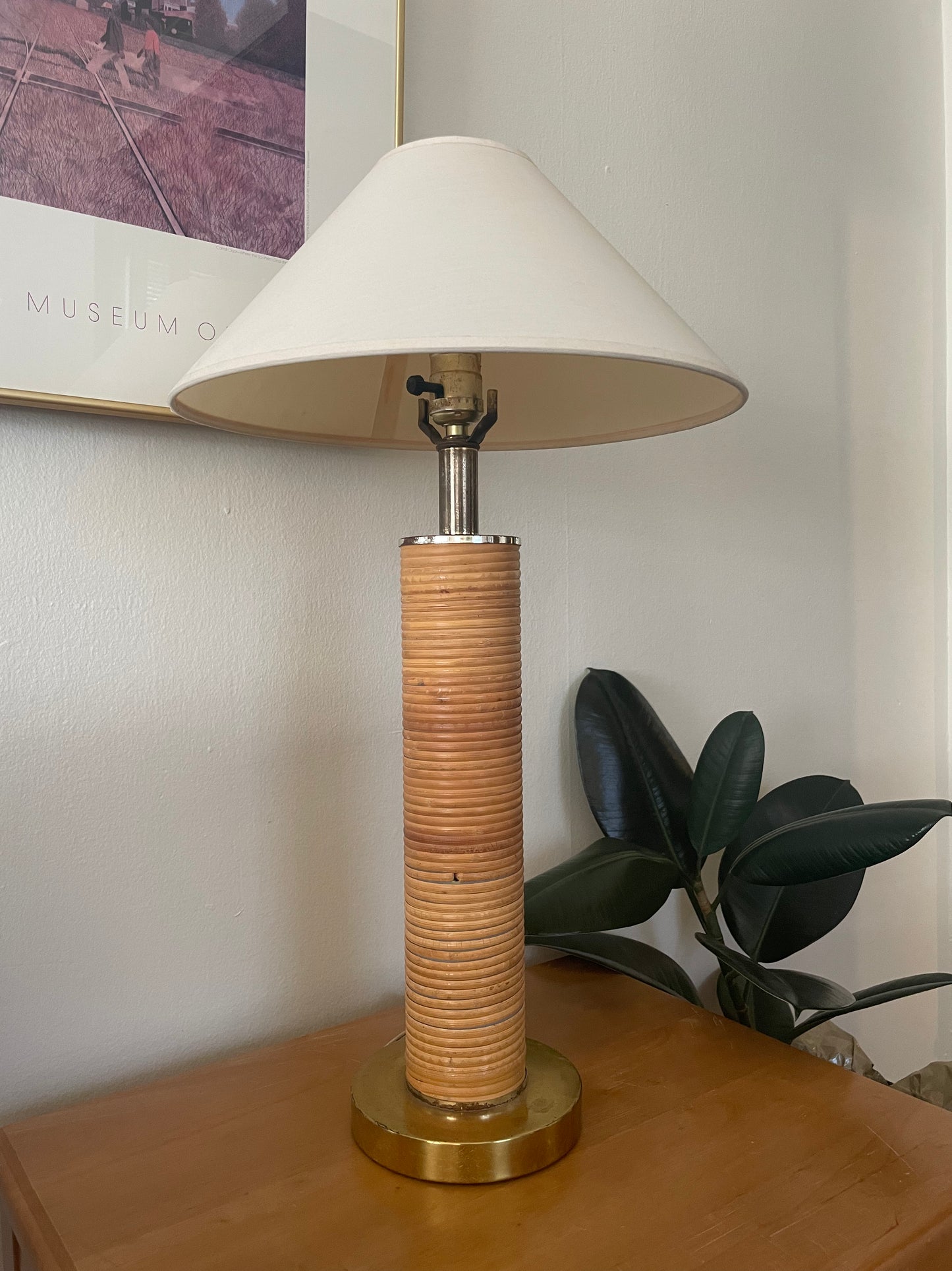 A table lamp with a cream linen taper shade, a silver metal neck, and slender cylindrical base wrapped in light rattan shown on a wood table next to a plant in front of a white wall with artwork.