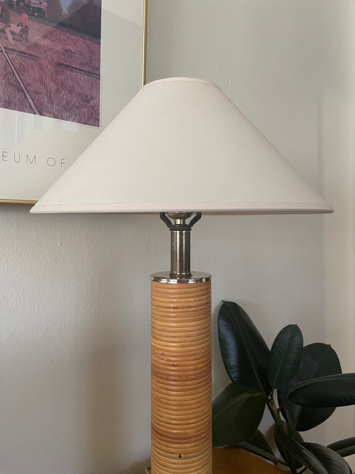 A table lamp with a cream linen taper shade, a silver metal neck, and slender cylindrical base wrapped in light rattan next to a plant and in front of artwork on a white wall.