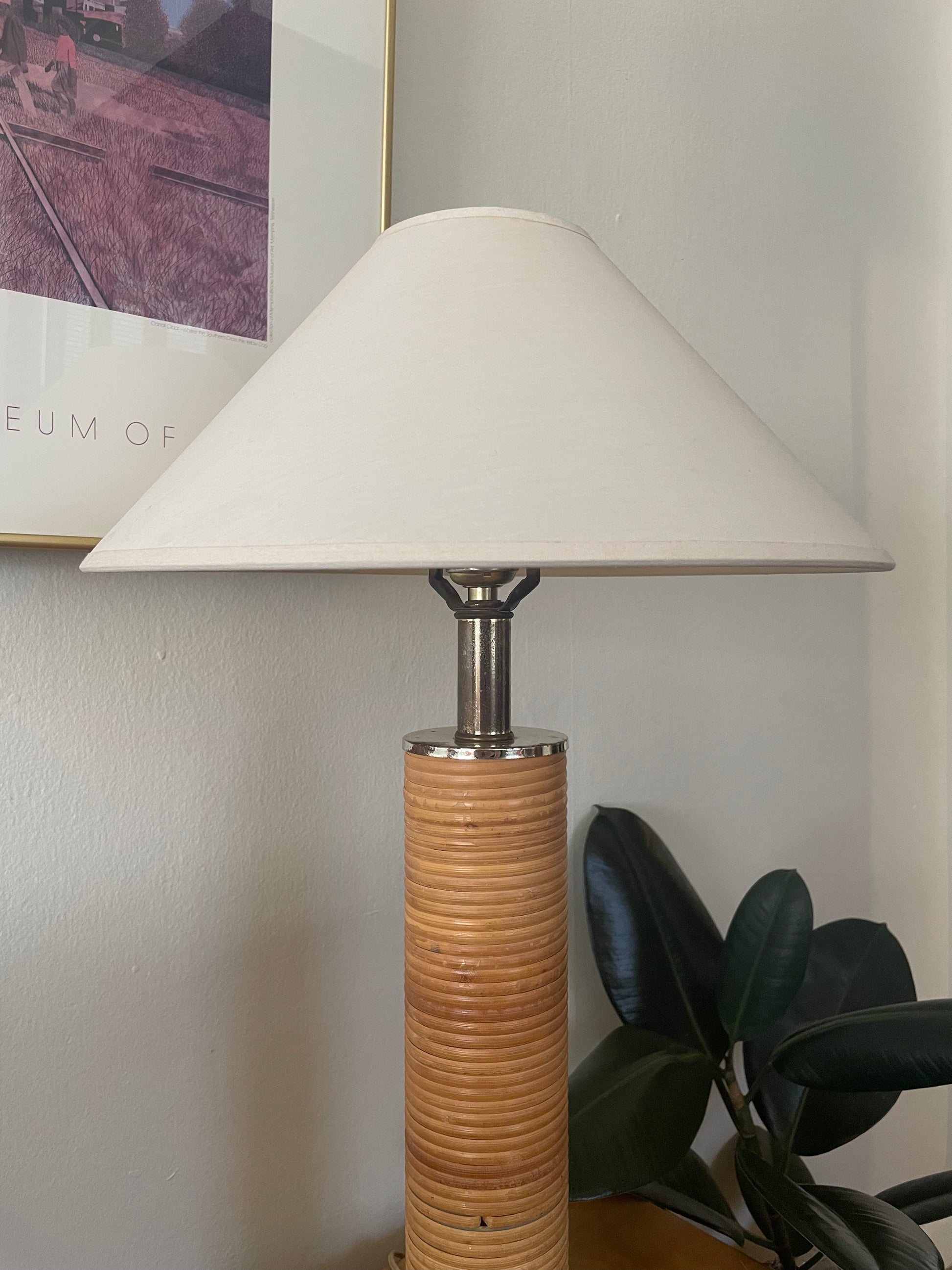A table lamp with a cream linen taper shade, a silver metal neck, and slender cylindrical base wrapped in light rattan next to a plant and in front of artwork on a white wall.