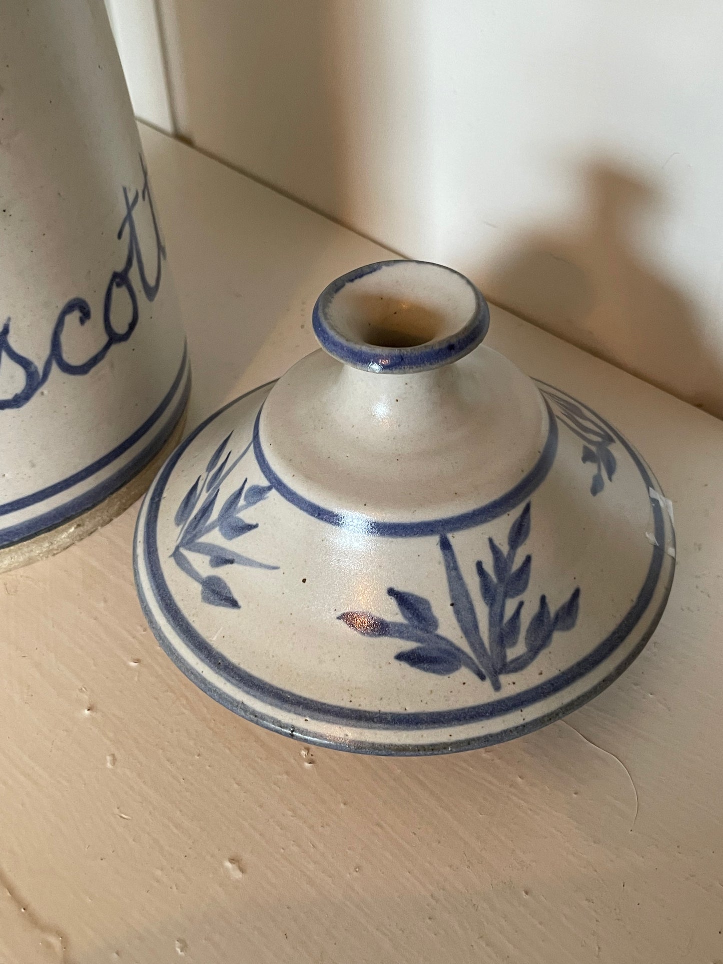 A small hand-painted ceramic lid with a blue floral design on an off-white glaze, resting on a light wooden surface next to a matching ceramic jar.