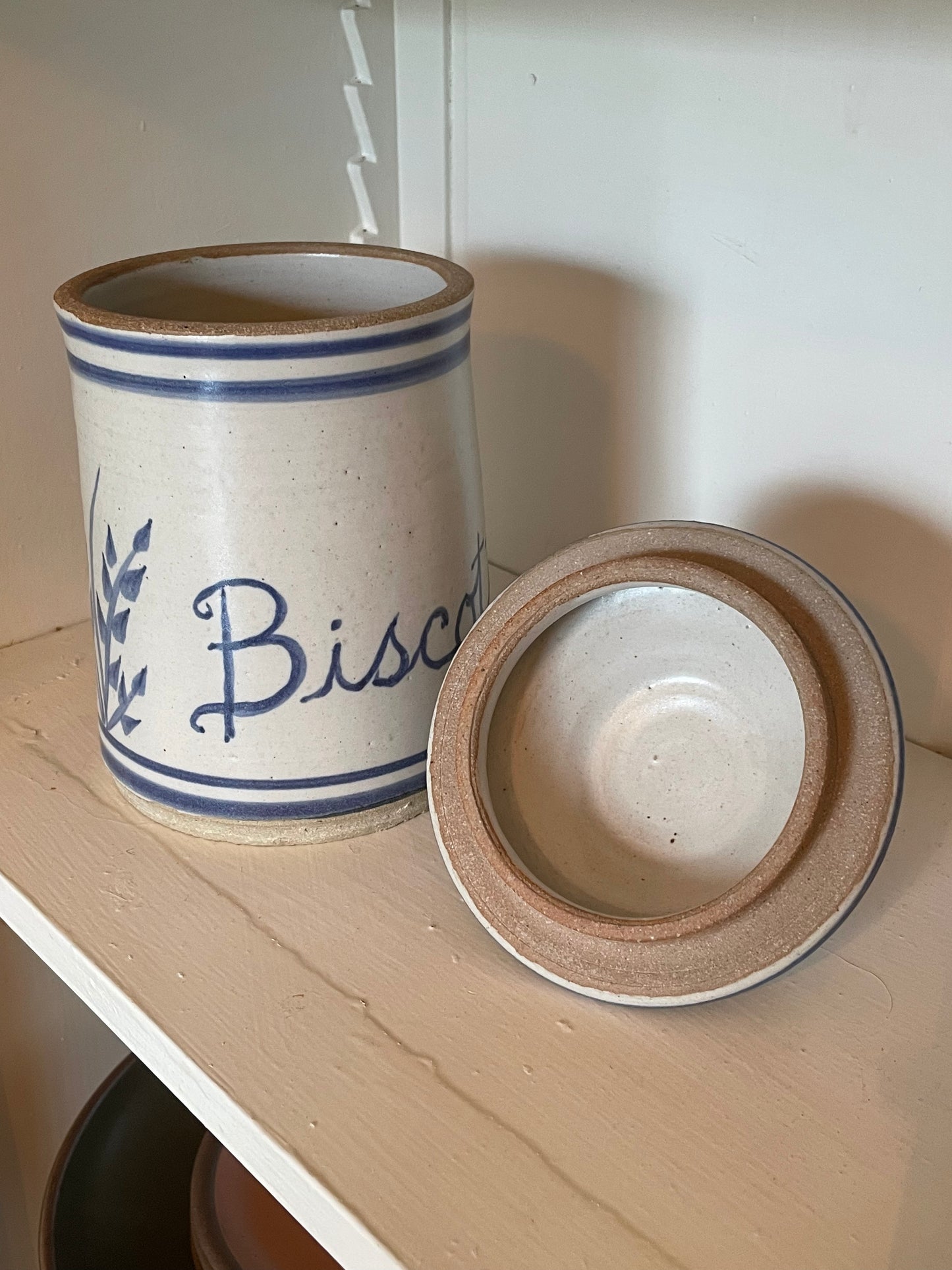 A rustic ceramic biscotti jar with a matching lid resting on a shelf. The jar is off-white with blue painted lettering spelling 'Biscotti' and blue decorative stripes, with a natural brown clay rim.