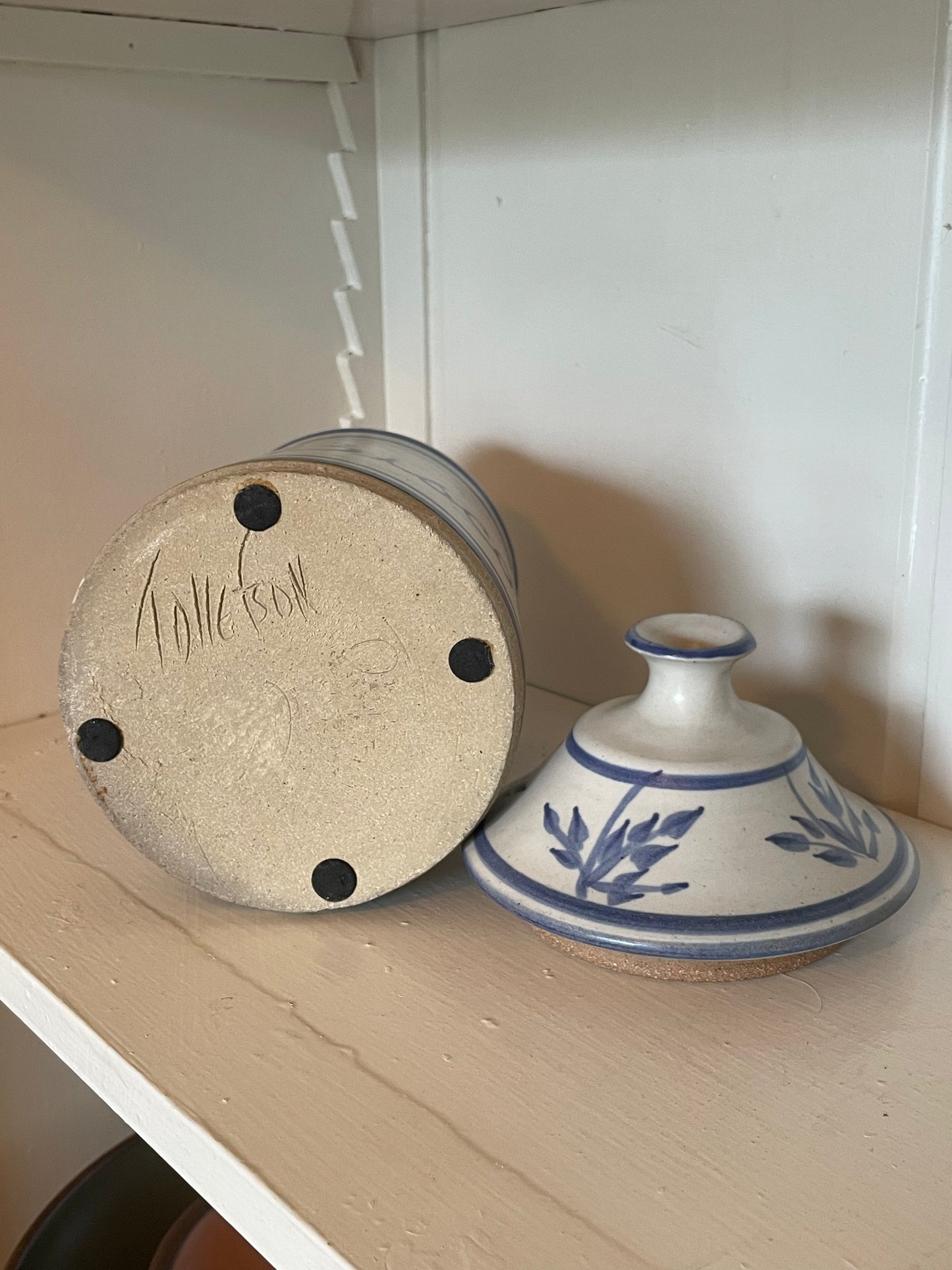 Ceramic lid with a blue floral pattern on a white glazed exterior, shown next to its upside-down beige unglazed clay base signed ‘Tollefson’ on a wooden shelf.
