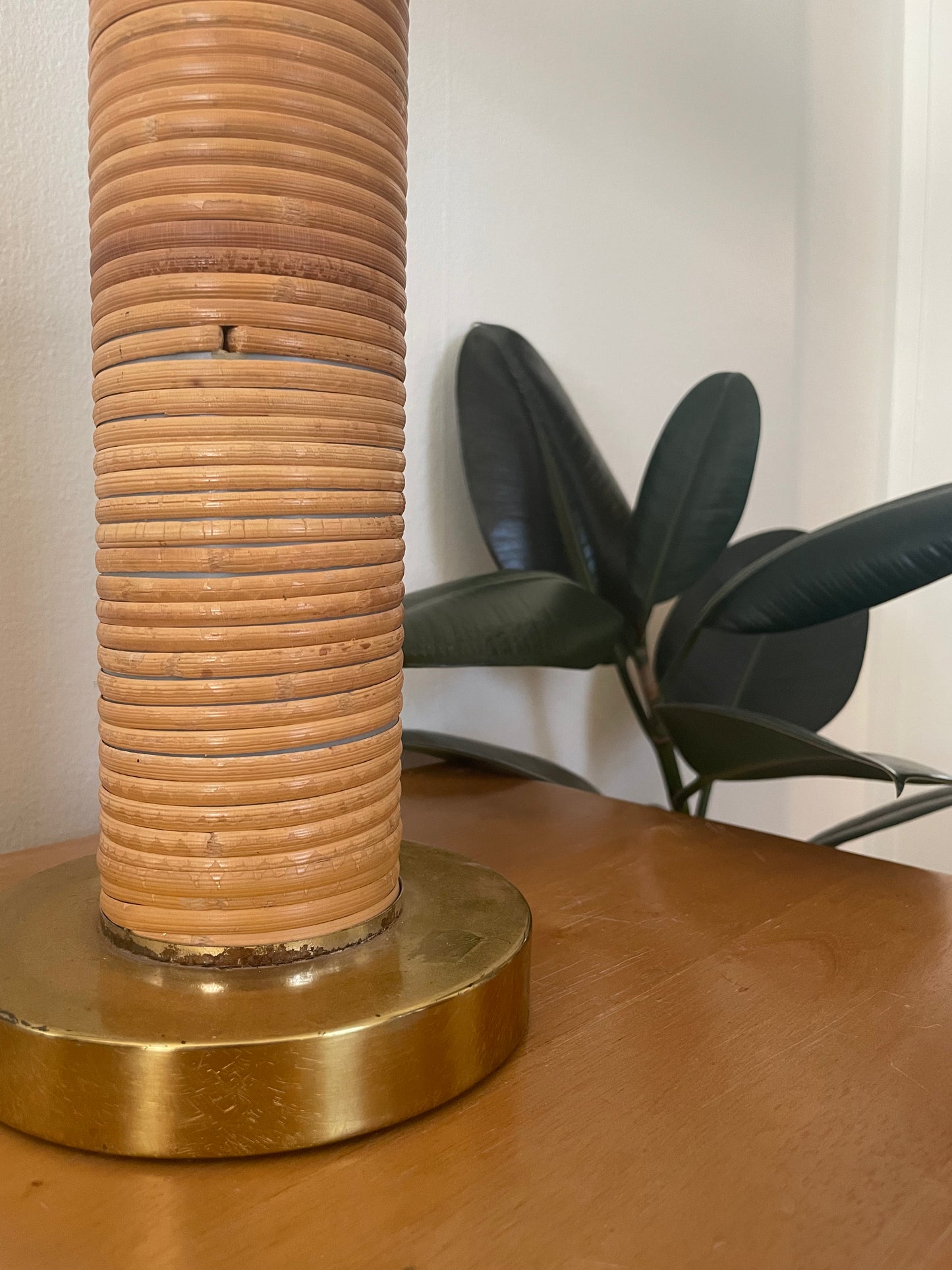 A close-up of a table lamp featuring a cylindrical column wrapped in light brown rattan on a round, polished brass base, set on a wooden table with a green plant in the background.
