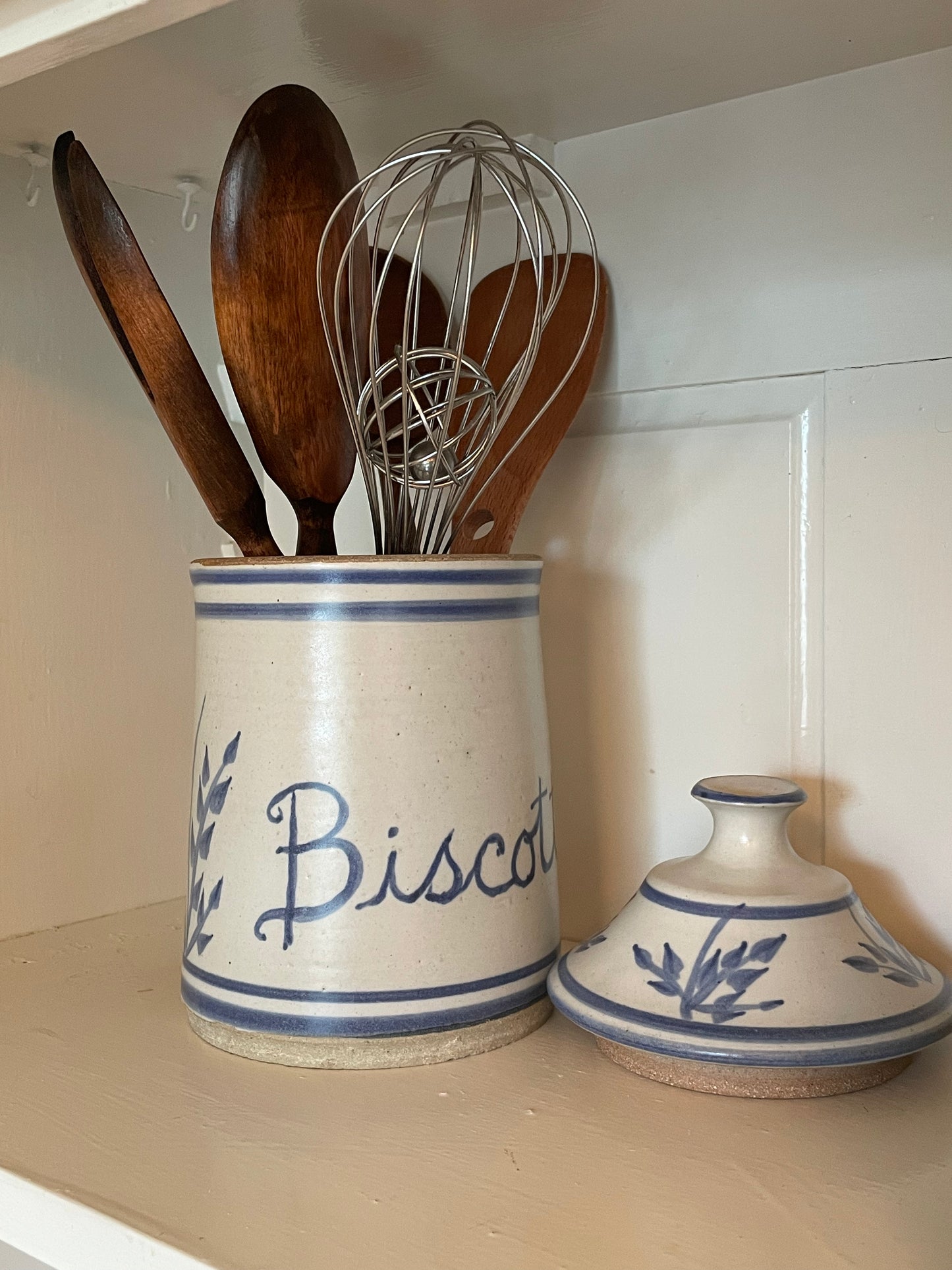 A ceramic kitchen utensil holder labeled 'Biscotti' in blue script, filled with wooden spoons and a metal whisk, displayed on a shelf next to a matching lid.