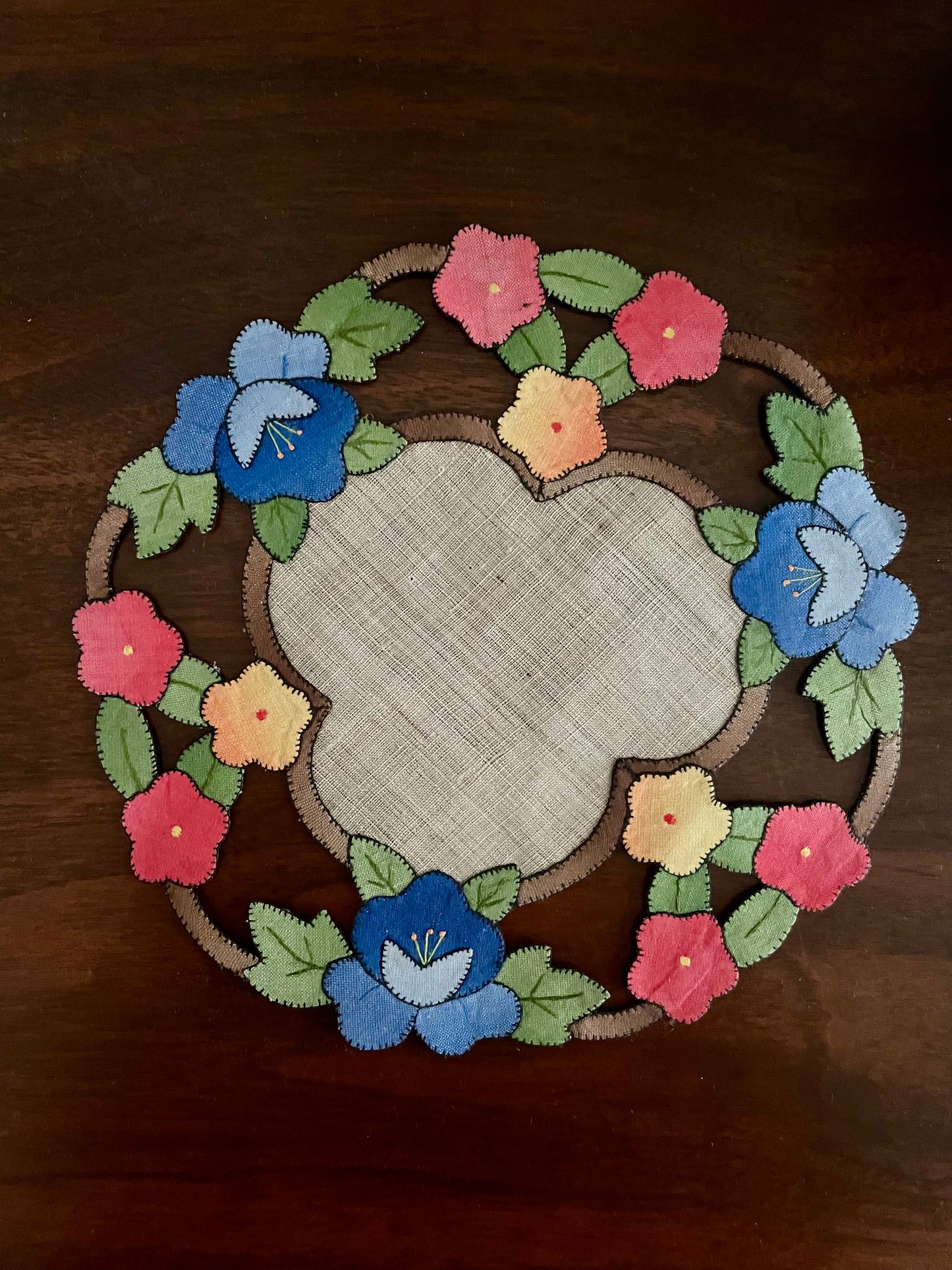 A round embroidered fabric placemat with a floral wreath design on a dark wooden table. The placemat features a central cream-colored linen-like circle surrounded by blue, pink, and yellow flowers with green leaves.