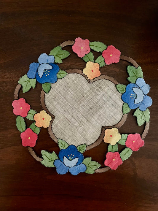 A round embroidered fabric placemat with a floral wreath design on a dark wooden table. The placemat features a central cream-colored linen-like circle surrounded by blue, pink, and yellow flowers with green leaves.