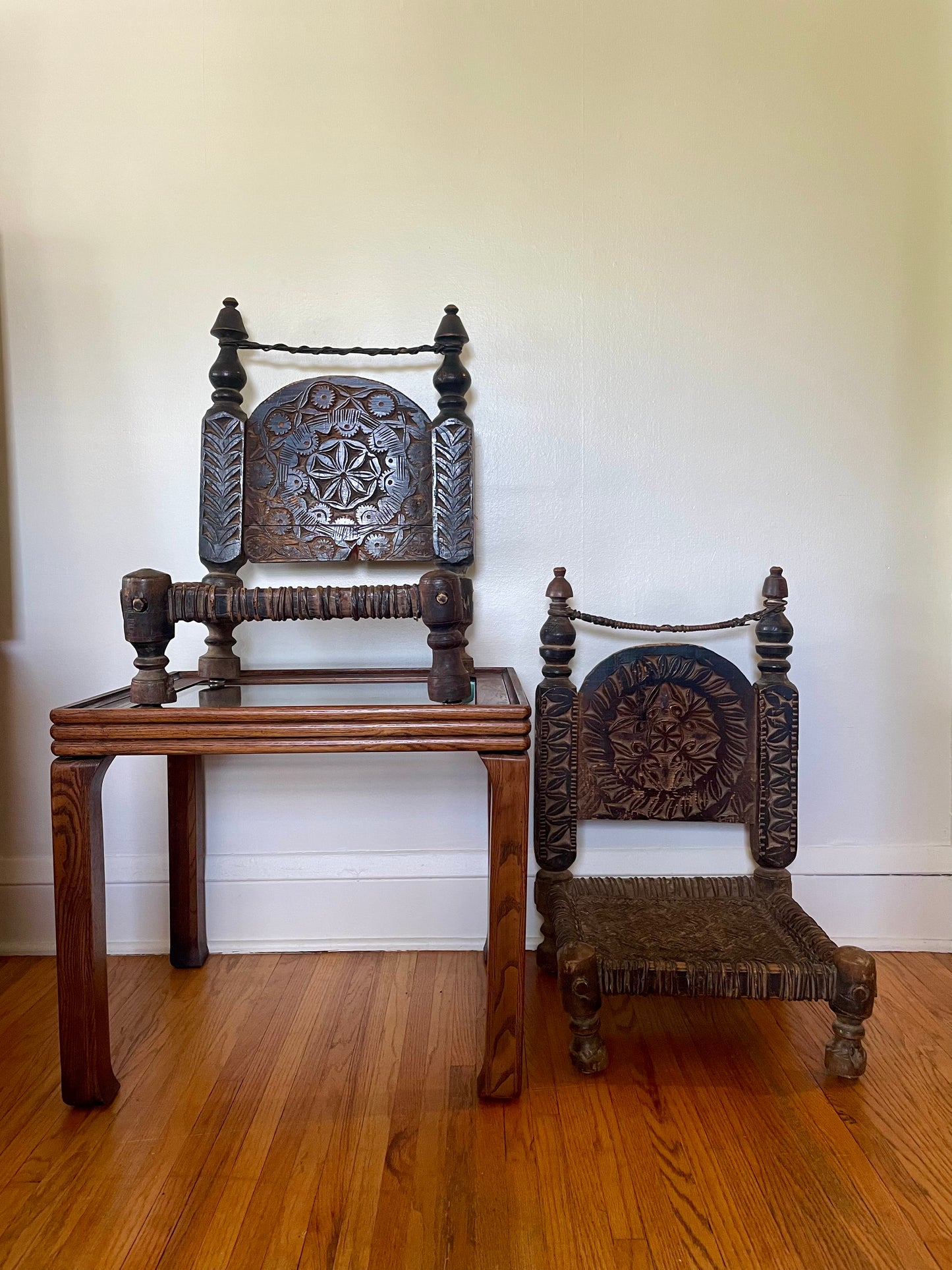Two dark carved wooden low chairs with ornately carved backrests. The chair on the right is sitting on the hardwood floor and the chair on the left is sitting on top of a small wooden side table, against a plain off-white wall.