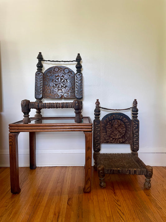 Two dark carved wooden low chairs with ornately carved backrests. The chair on the right is sitting on the hardwood floor and the chair on the left is sitting on top of a small wooden side table, against a plain off-white wall.
