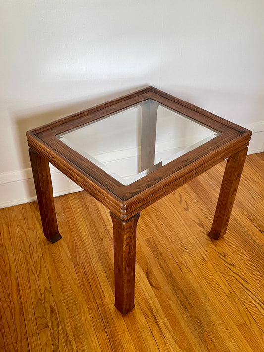A rectangular wooden end table with a clear glass top, featuring a walnut brown wood frame and four legs, set on a hardwood floor against a white wall.