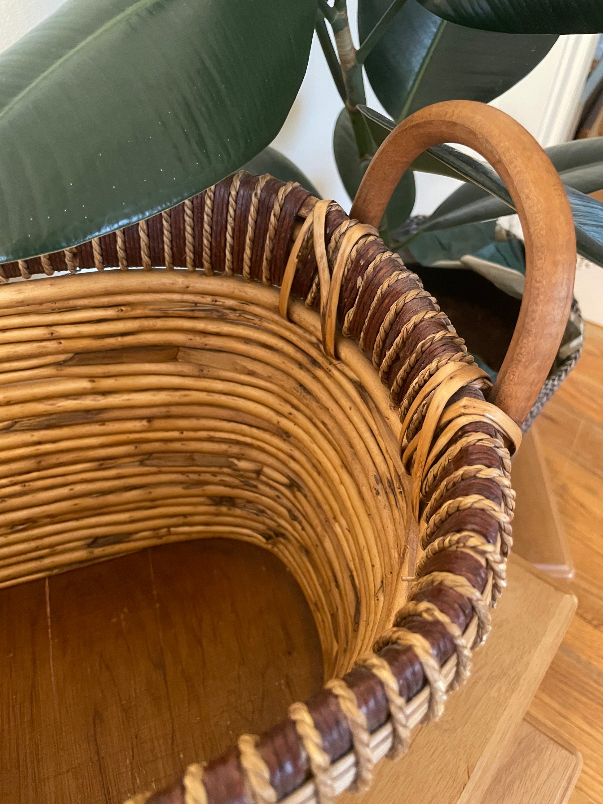 A close-up of a woven rattan storage basket with a curved bentwood handle, set on a wooden surface next to a potted rubber plant.