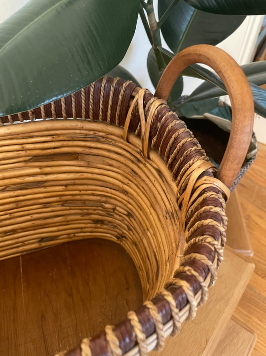 A close-up of a woven rattan storage basket with a curved bentwood handle, set on a wooden surface next to a potted rubber plant.