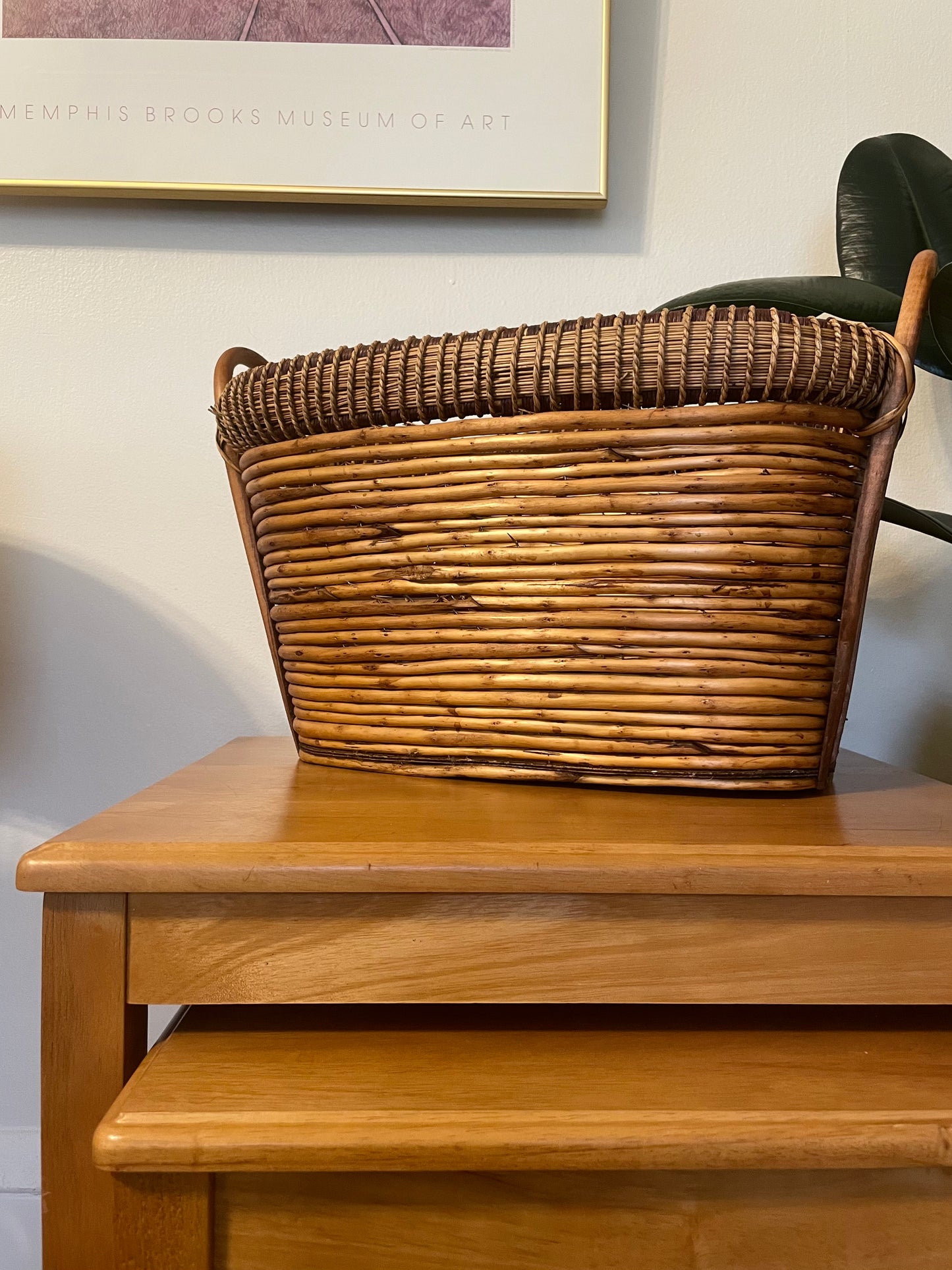 A brown woven rattan basket with a long oval shape and curved bentwood handles, sitting on a wooden side table in an indoor setting.