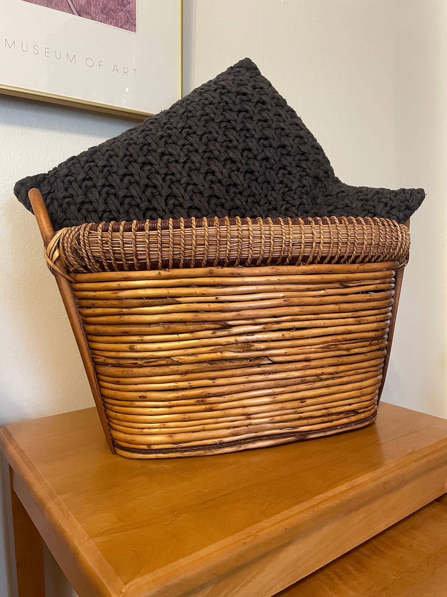 A charcoal grey knit throw pillow resting inside a woven rattan basket on a wooden table, with a cream wall and framed art in the background.
