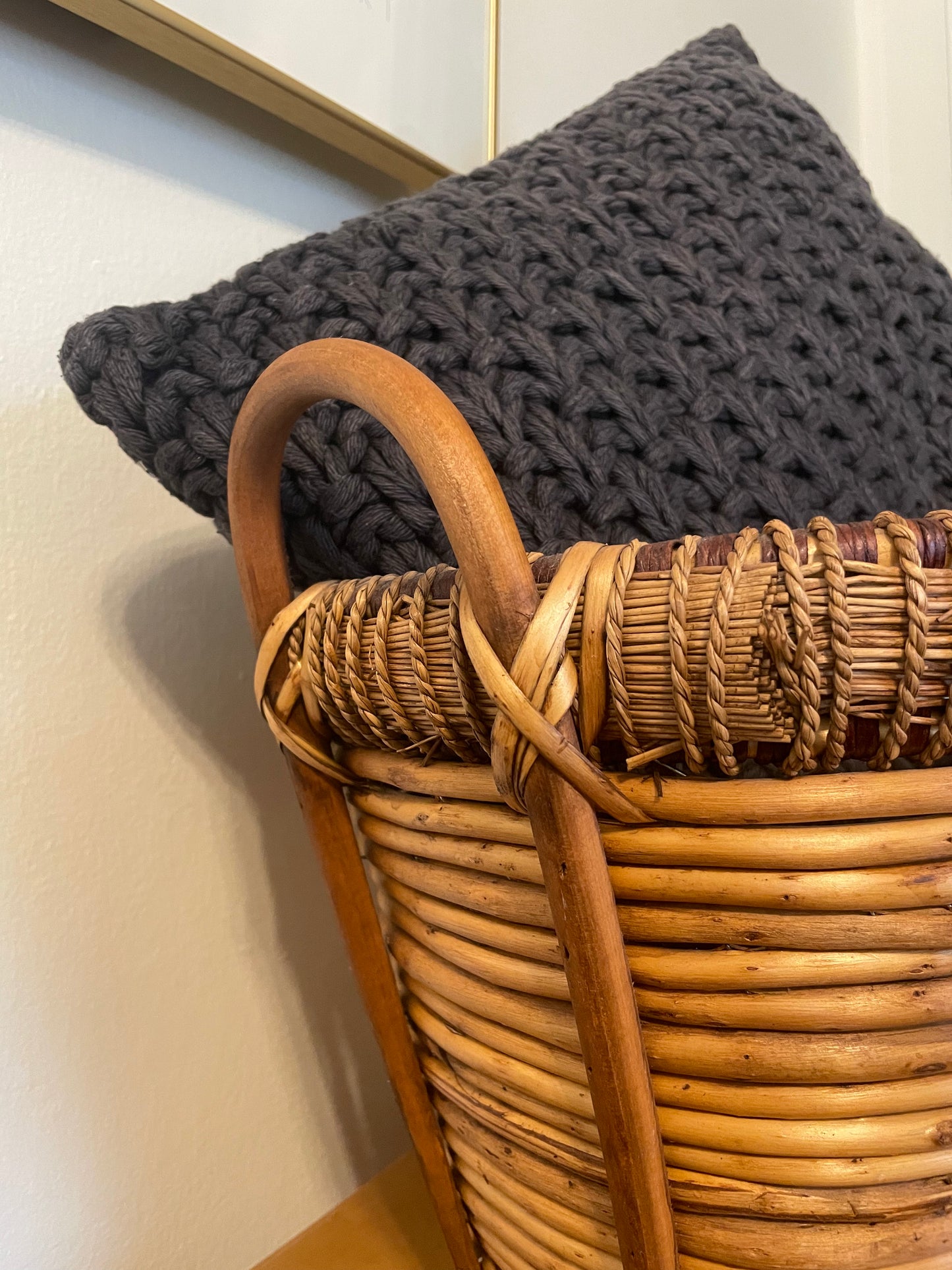 A close-up of a dark charcoal gray chunky knit throw pillow resting inside a natural woven rattan basket with a wooden handle, set against a neutral beige wall.