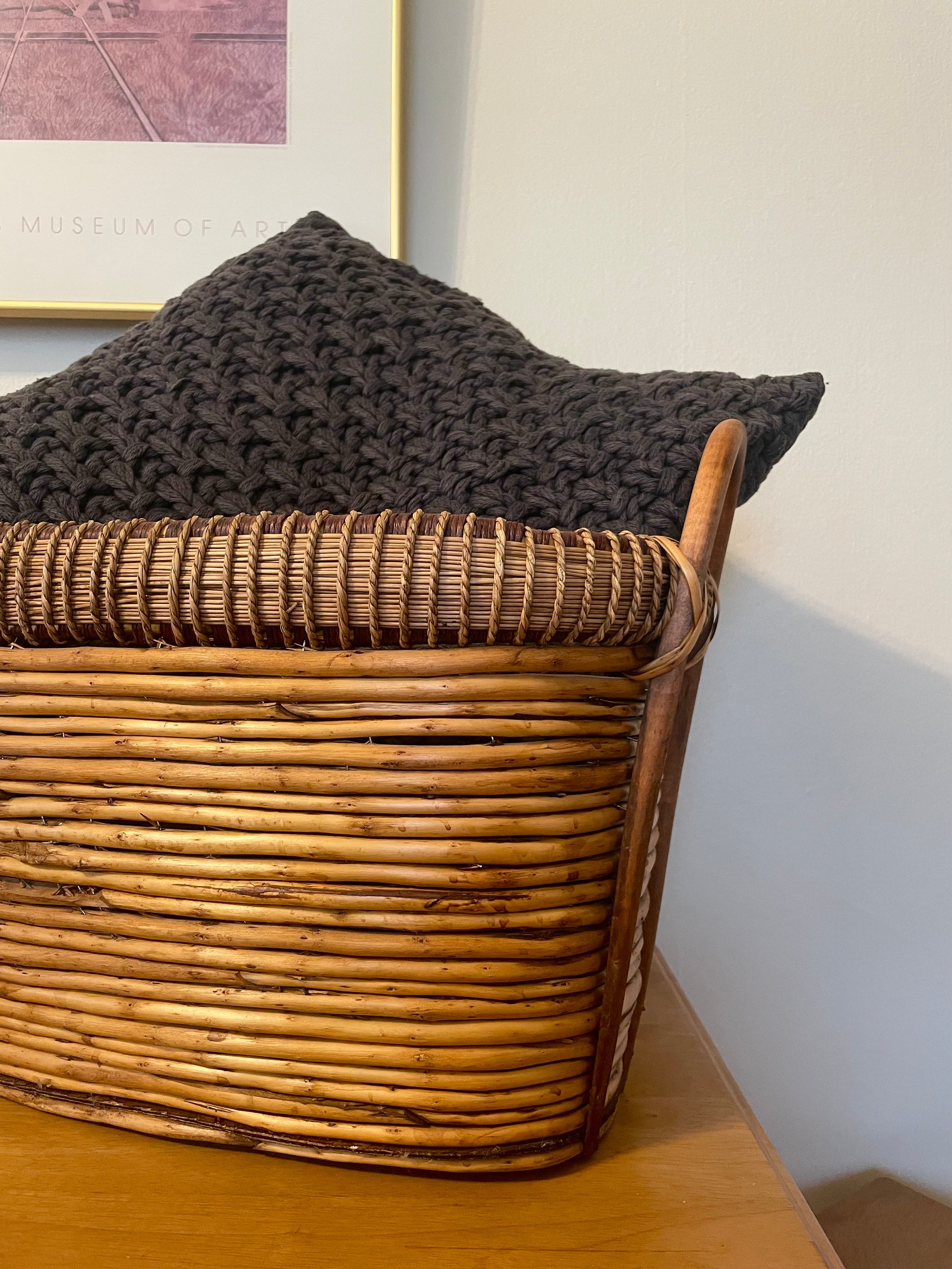 A close-up of a natural woven rattan storage basket with a dark charcoal knit throw pillow resting inside, set on a wooden table against a white wall.