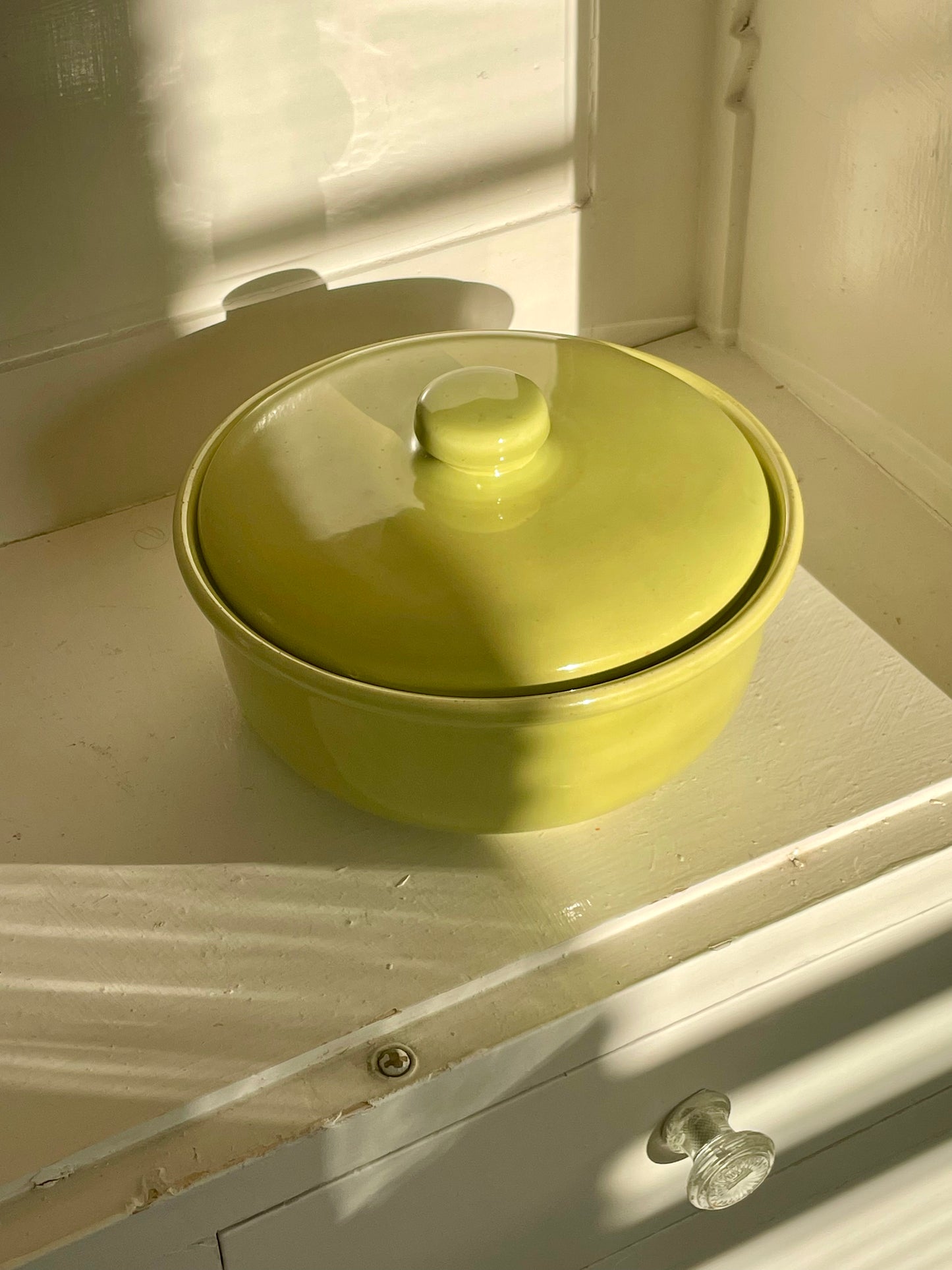 A bright yellow-green ceramic lidded casserole dish sits inside of a white cabinet with warm shadows falling across the piece.