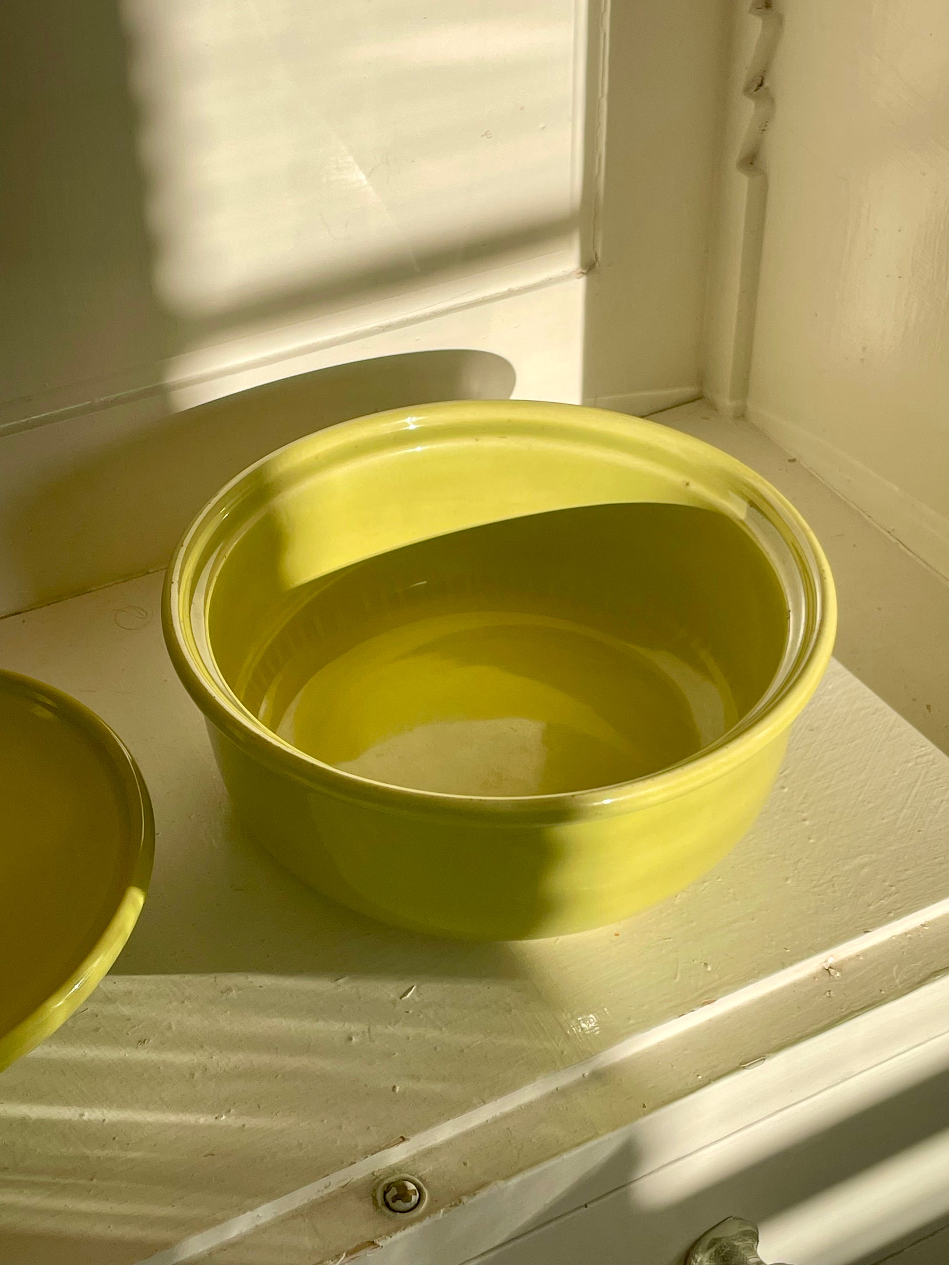 A bright yellow-green ceramic casserole dish sits inside of a white cabinet with warm shadows falling across the piece and the matching lid partially shown at the edge of the image.