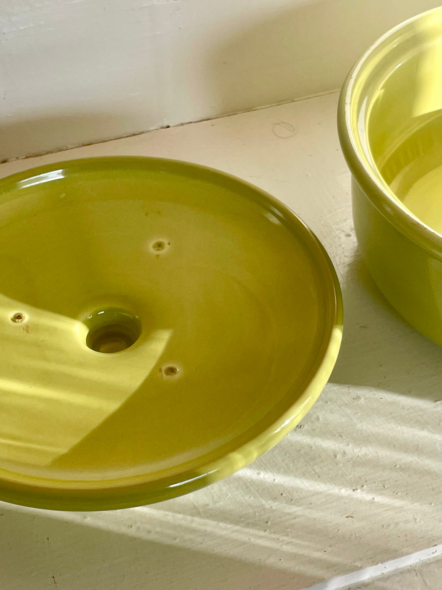 Closeup of the underside of a bright yellow-green ceramic lid sitting inside of a white cabinet with warm shadows falling across it. The matching casserole dish is partially shown at the edge of the image.
