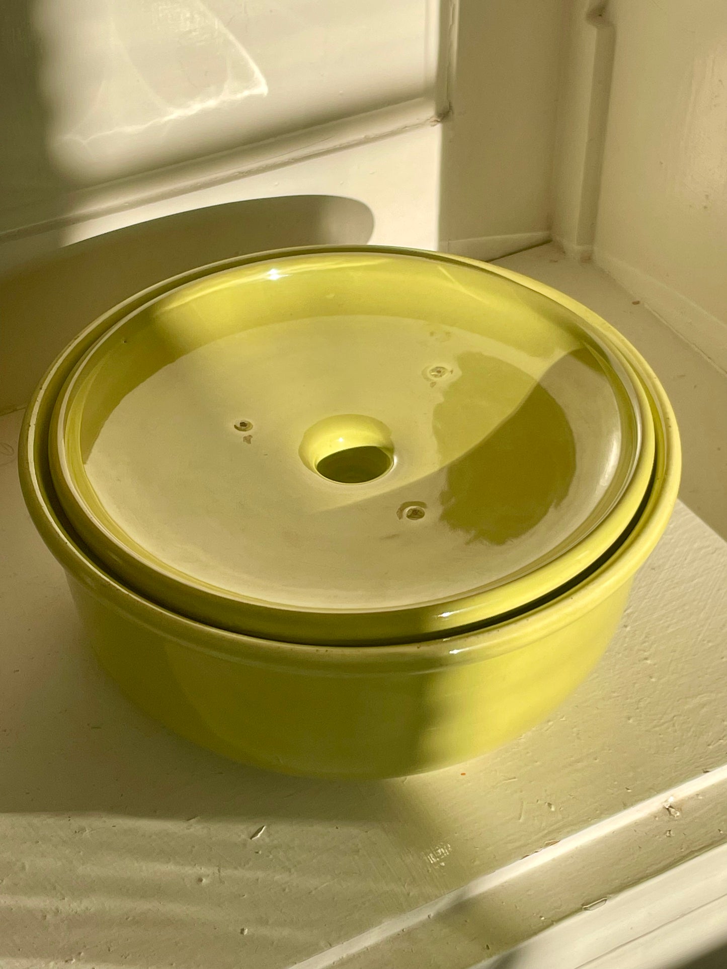 A bright yellow-green ceramic lidded casserole dish sits inside of a white cabinet with warm shadows falling across the piece. Its lid is show resting upside-down inside of the dish.