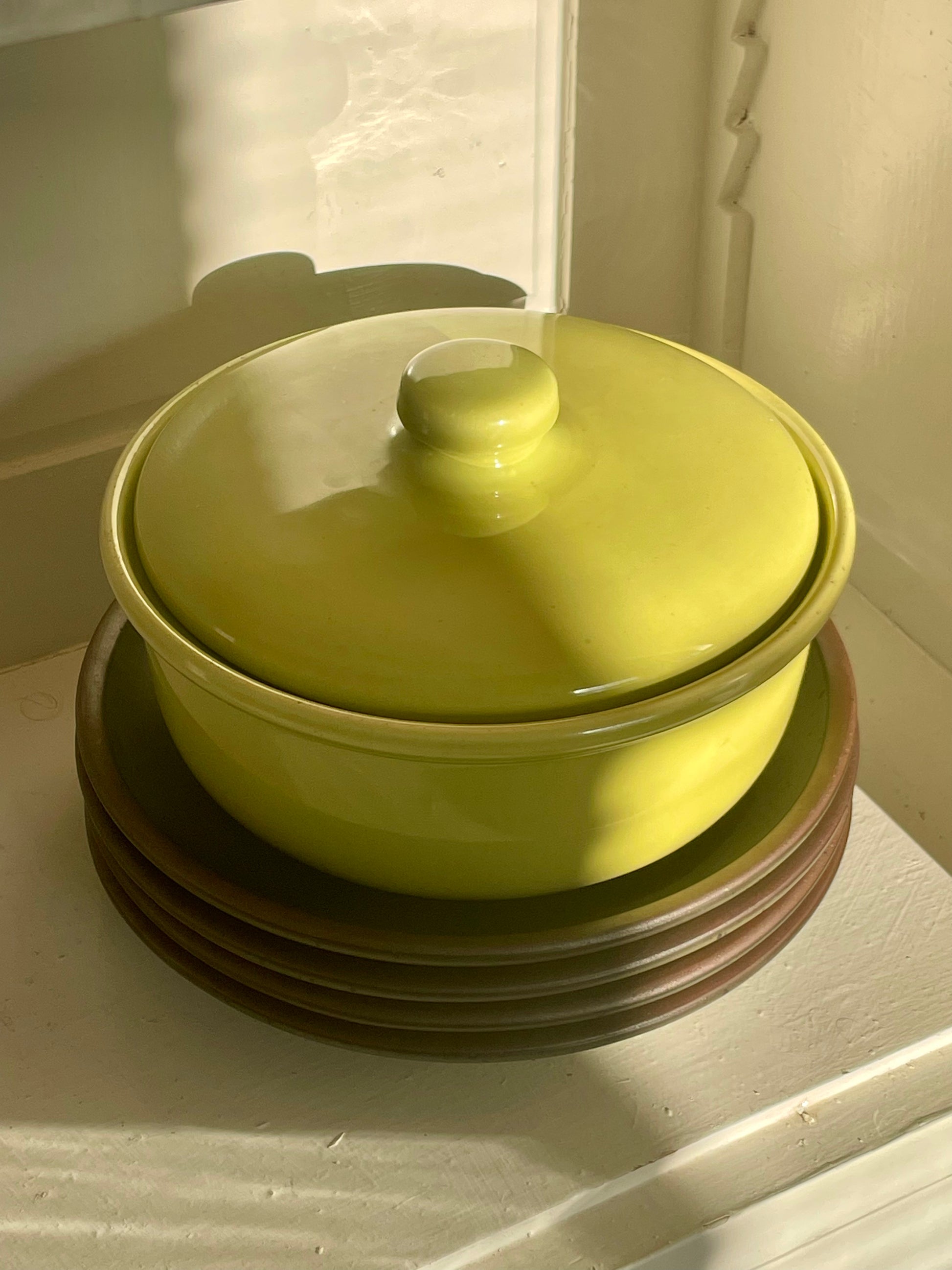 A bright yellow-green ceramic lidded casserole dish sits on a stack of dark green plates with raw clay edges inside of a white cabinet, lit by warm natural light with shadows falling across the piece.