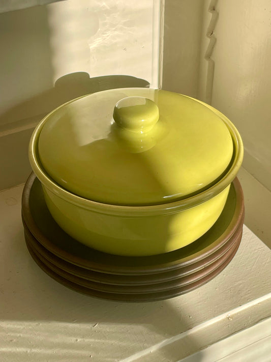 A bright yellow-green ceramic lidded casserole dish sits on a stack of dark green plates with raw clay edges inside of a white cabinet, lit by warm natural light with shadows falling across the piece.