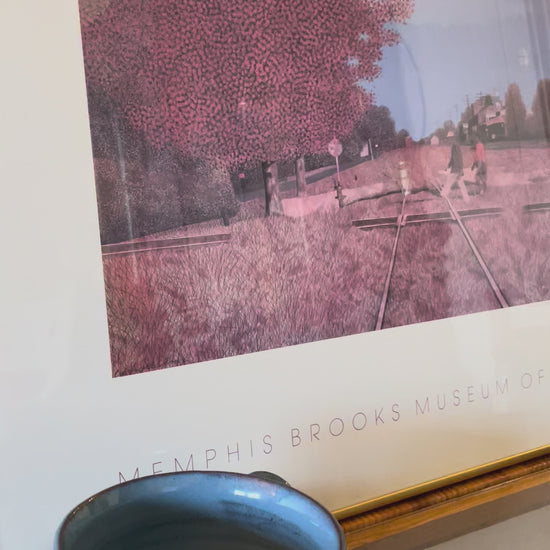 Video panning a framed art print from the Memphis Brooks Museum of Art, featuring a stylized railroad scene with a large pinkish-brown tree, two figures walking across the tracks, and a blue sky, mounted in a gold frame and leaning against a white wall.