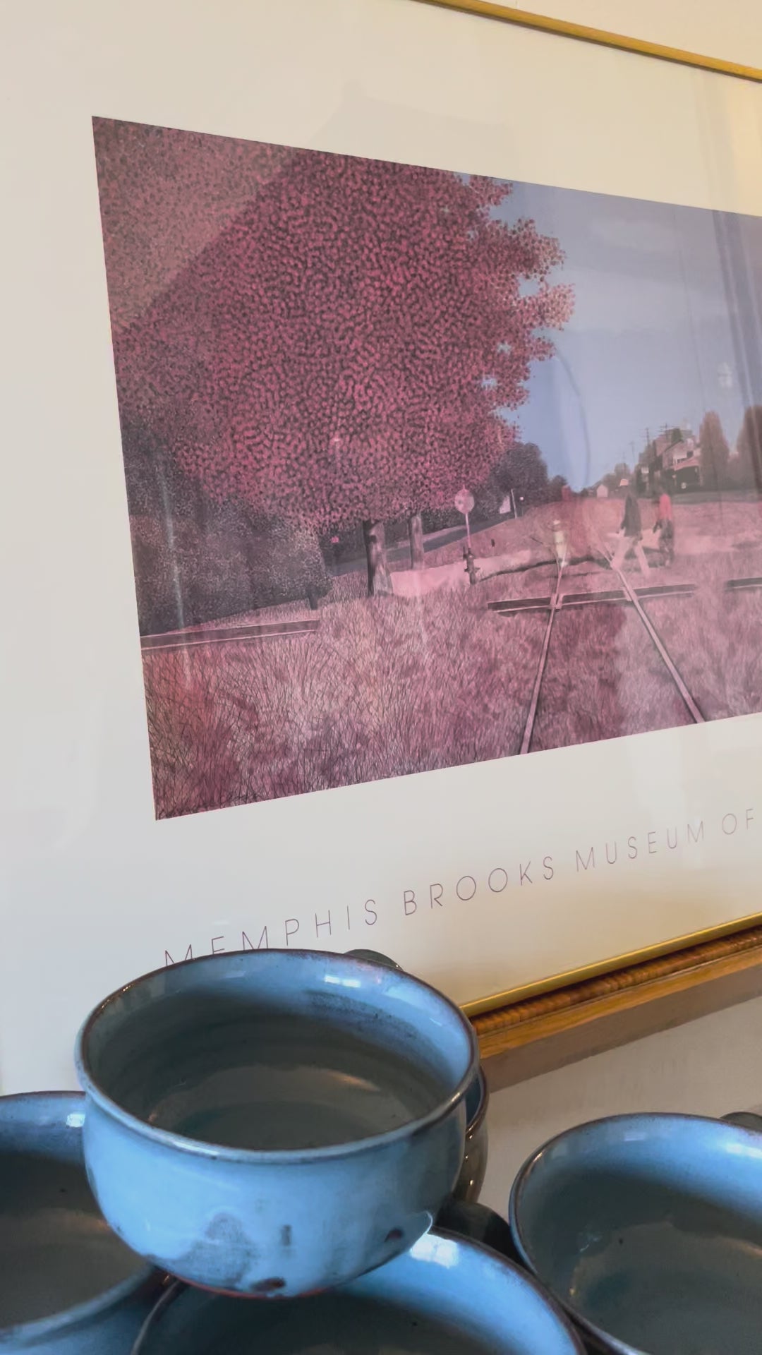 Video panning a framed art print from the Memphis Brooks Museum of Art, featuring a stylized railroad scene with a large pinkish-brown tree, two figures walking across the tracks, and a blue sky, mounted in a gold frame and leaning against a white wall.