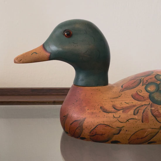 Video showing the swiveling head of a hand-painted duck decoy as it rests on a glass-top wood table. The duck decoy features a dark green head, orange-brown beak, and orange-brown body with painted botanical details.
