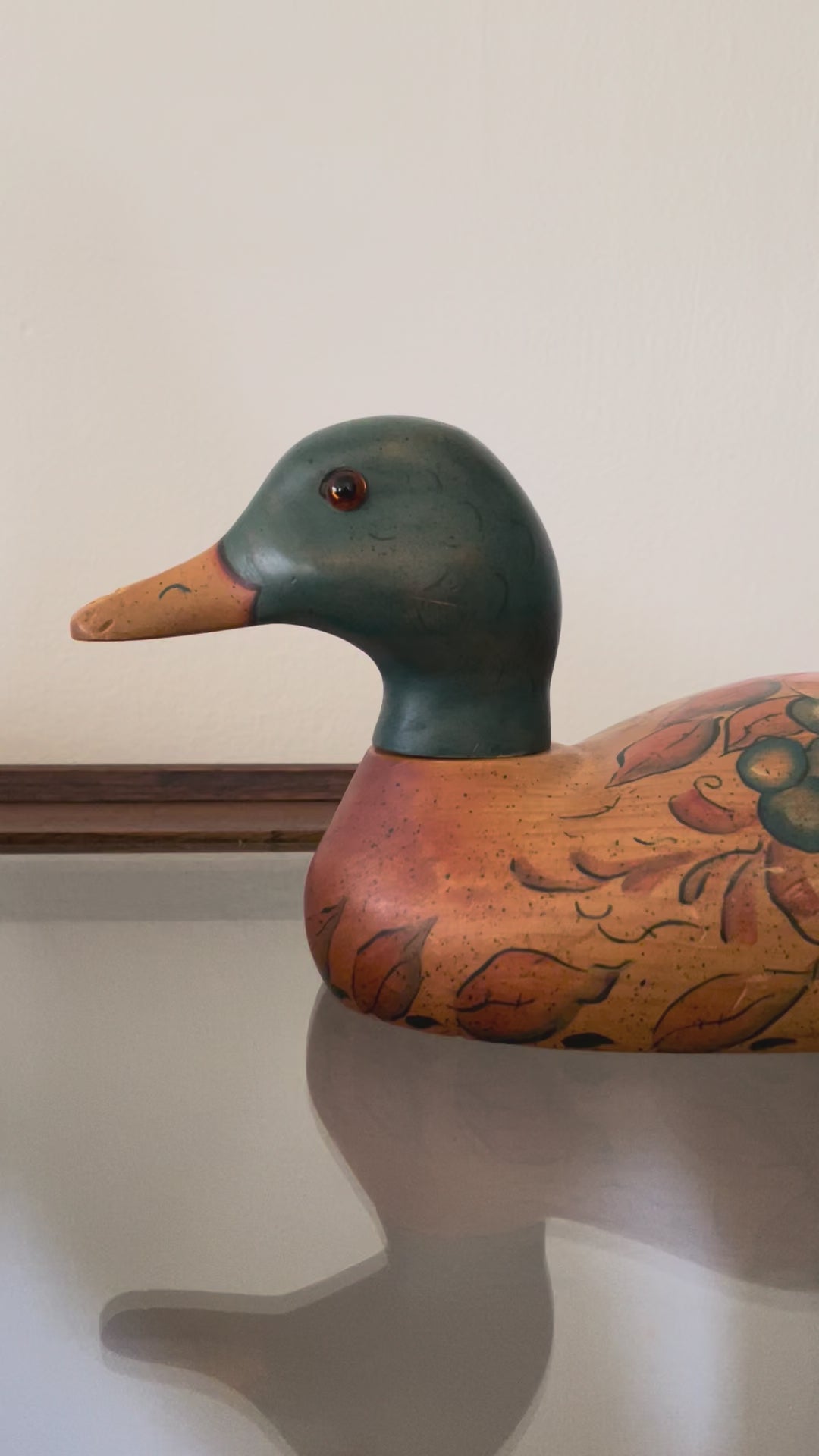 Video showing the swiveling head of a hand-painted duck decoy as it rests on a glass-top wood table. The duck decoy features a dark green head, orange-brown beak, and orange-brown body with painted botanical details.