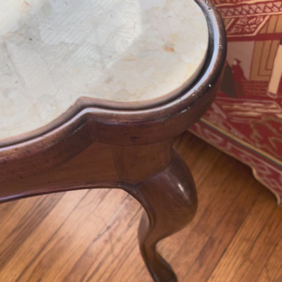 Video showing closeup pan of a cream marble-top table with scalloped wood frame, resting on wood floors in front of a white wall.