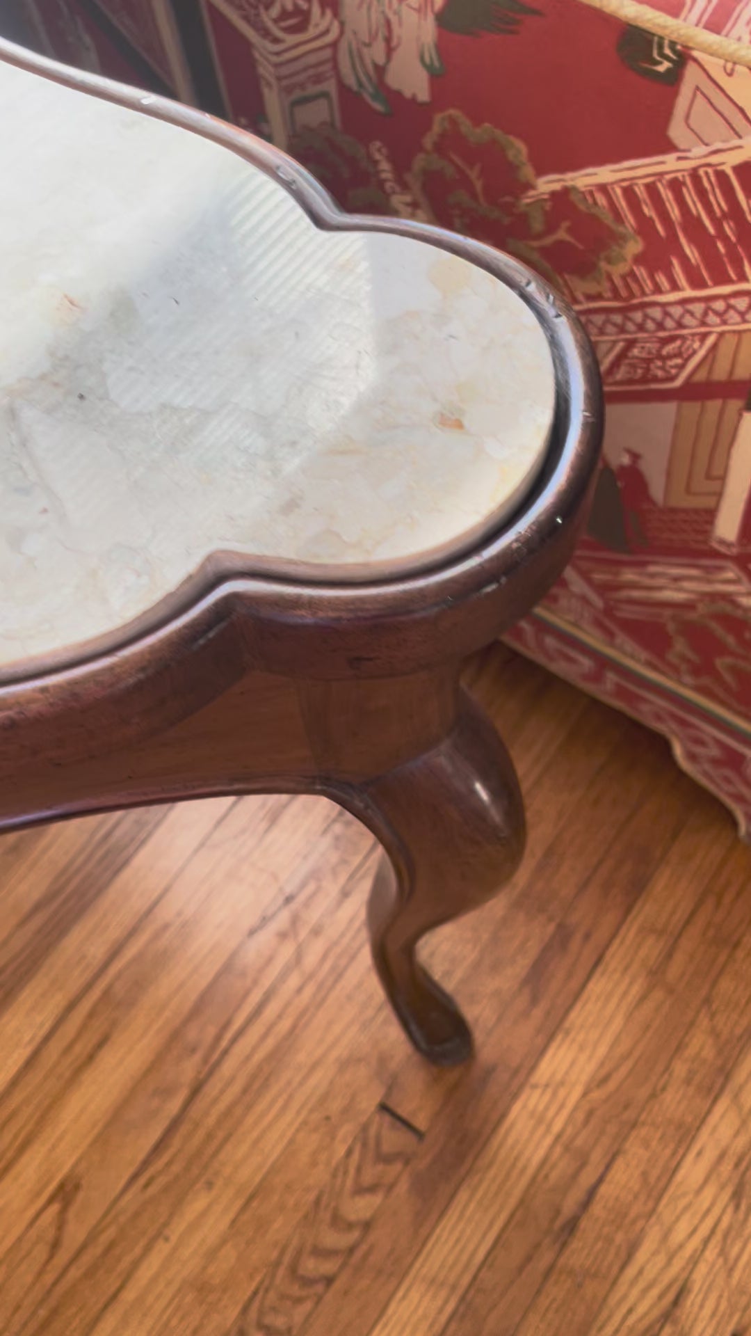 Video showing closeup pan of a cream marble-top table with scalloped wood frame, resting on wood floors in front of a white wall.