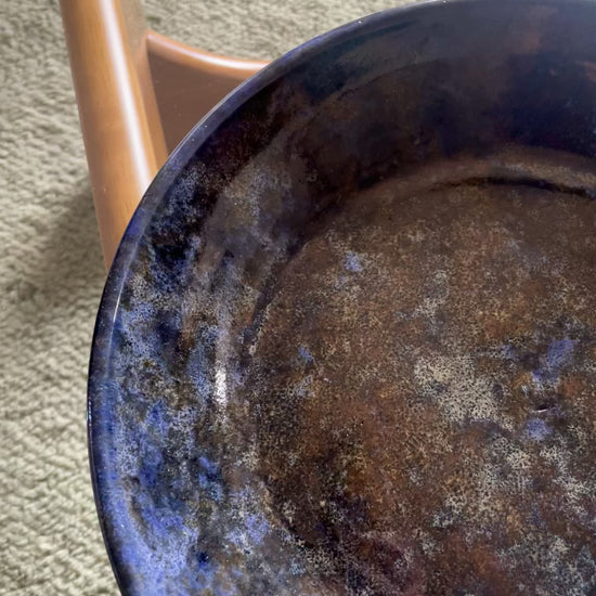 A video shows a close-up pan of a large round ceramic bowl with speckled blue and brown glazing rests on a glass table over a blue-grey rug.