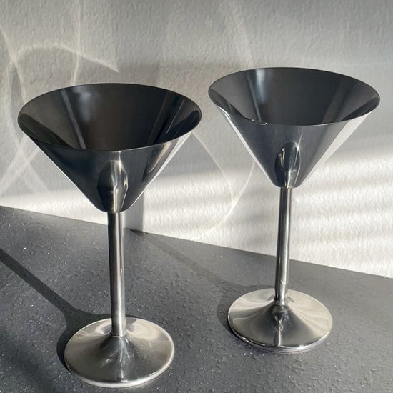 Video of two polished silver metal cocktail muddlers with wide conical heads and slender stems, standing on a dark gray surface against a textured light gray wall with dramatic shadows dancing across them.