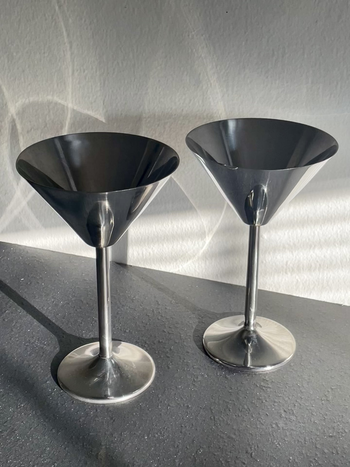 Video of two polished silver metal cocktail muddlers with wide conical heads and slender stems, standing on a dark gray surface against a textured light gray wall with dramatic shadows dancing across them.