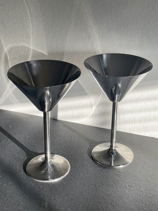 Video of two polished silver metal cocktail muddlers with wide conical heads and slender stems, standing on a dark gray surface against a textured light gray wall with dramatic shadows dancing across them.
